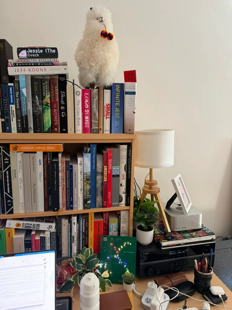 A packed bookshelf topped with a stuffed alpaca, beside a lamp.